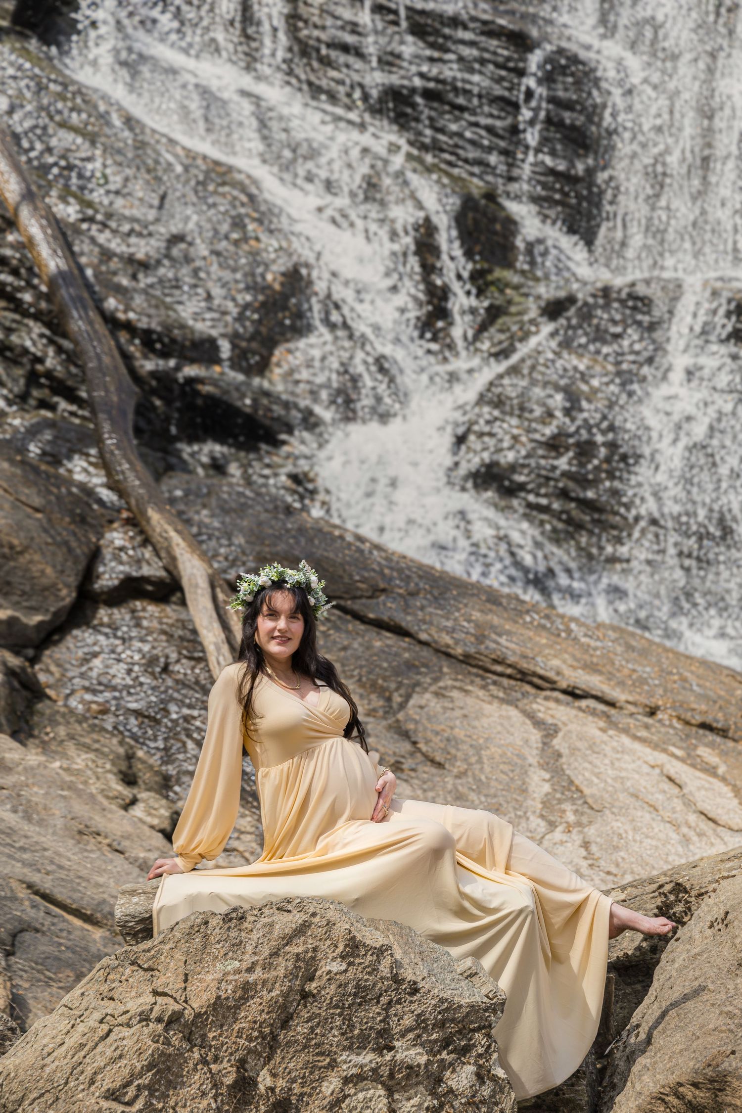Maternity photoshoot at scenic waterfall with woman in flowing cream dress wearing floral crown.