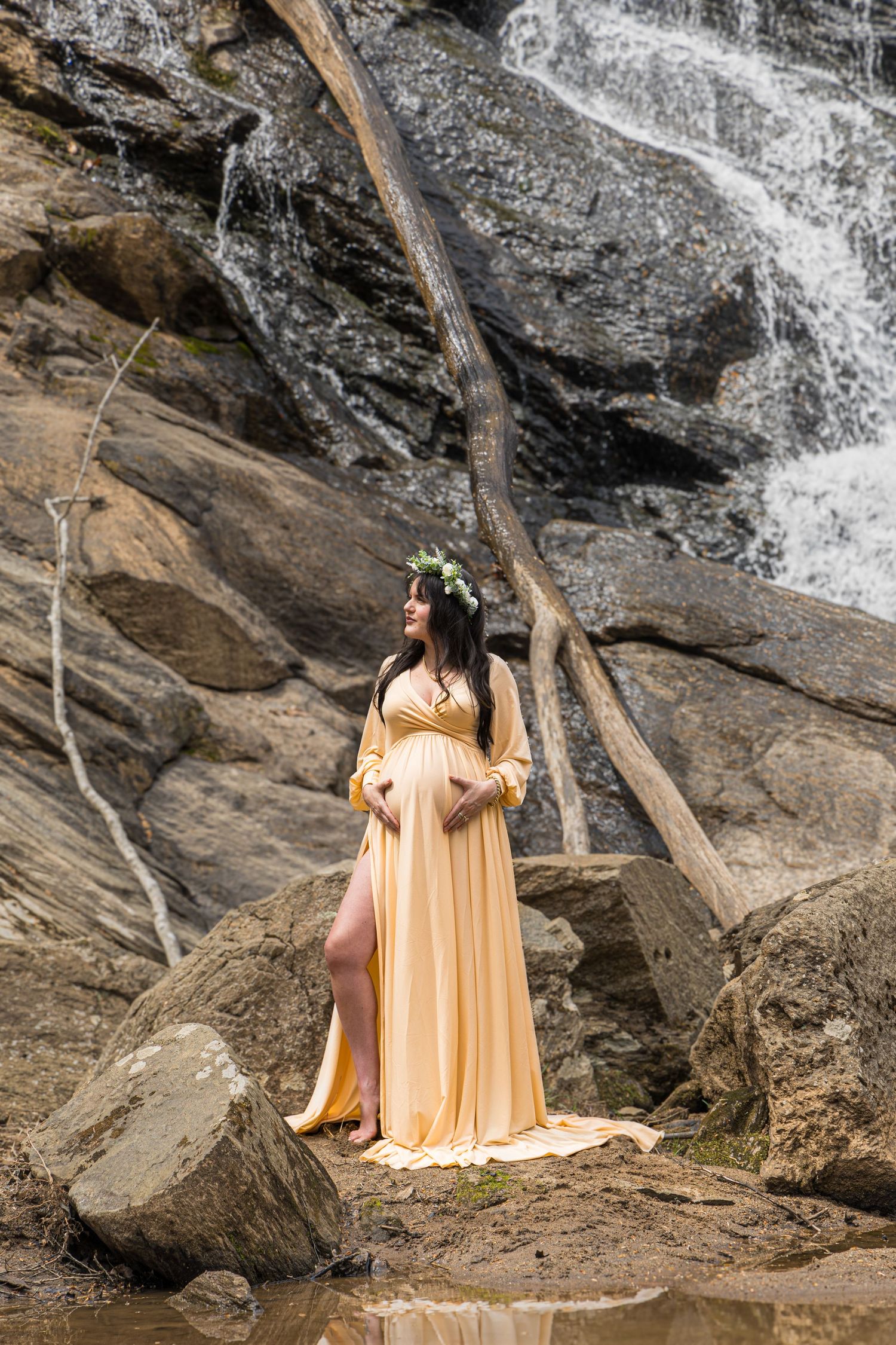 Maternity photo featuring woman in golden dress posing elegantly by rocky waterfall.