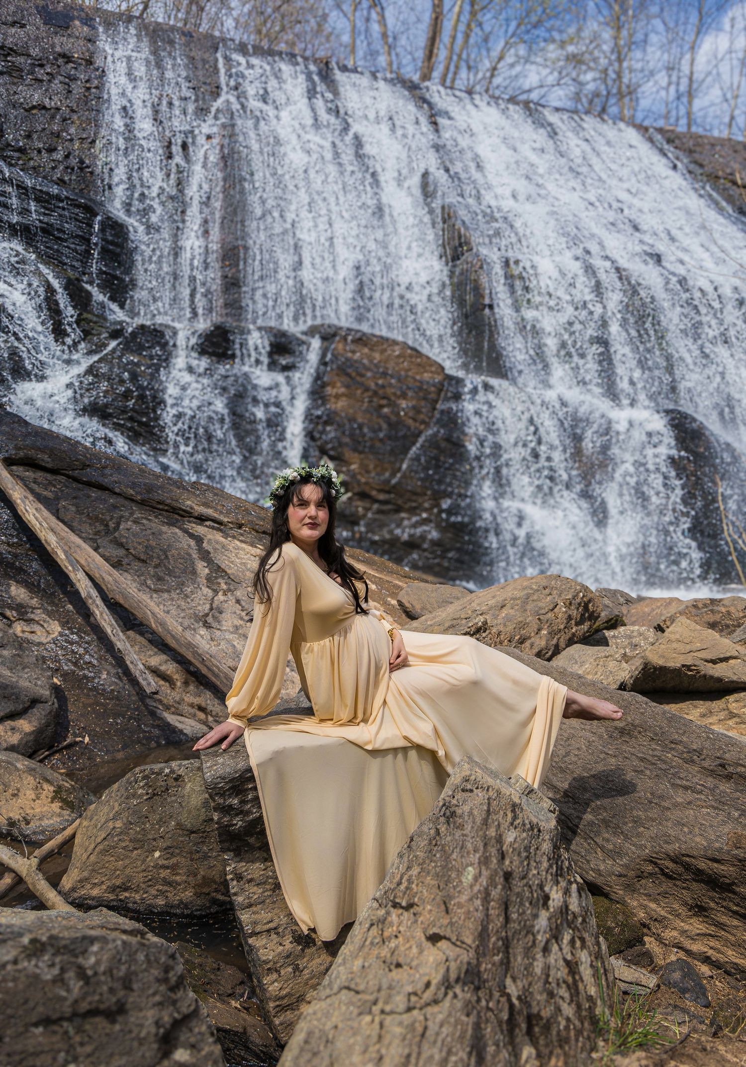 Expectant mother in flowing cream dress poses gracefully on rocks near rushing waterfall.