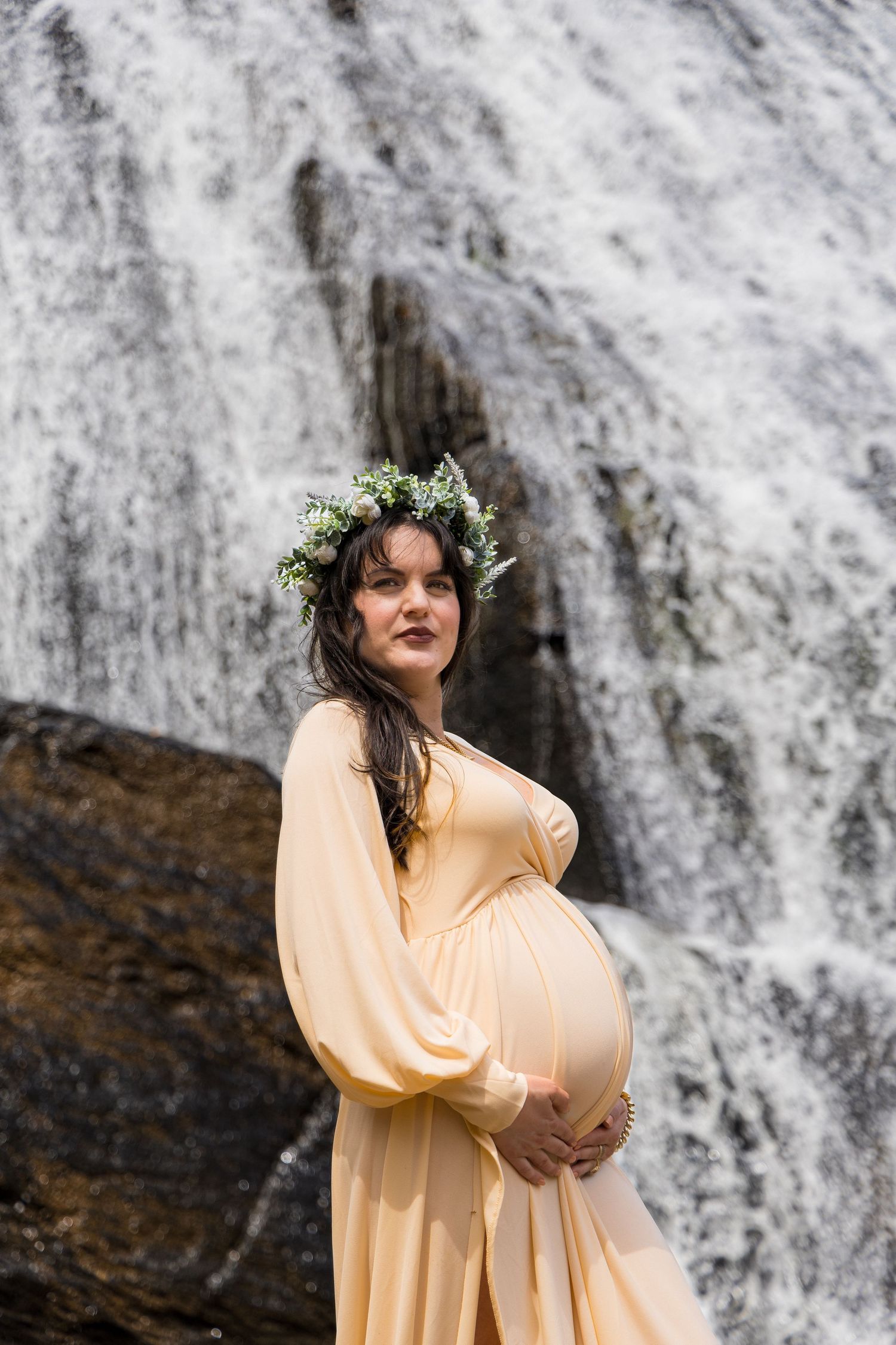 A person in a flowing peach dress with a floral crown stands before a cascading waterfall.
