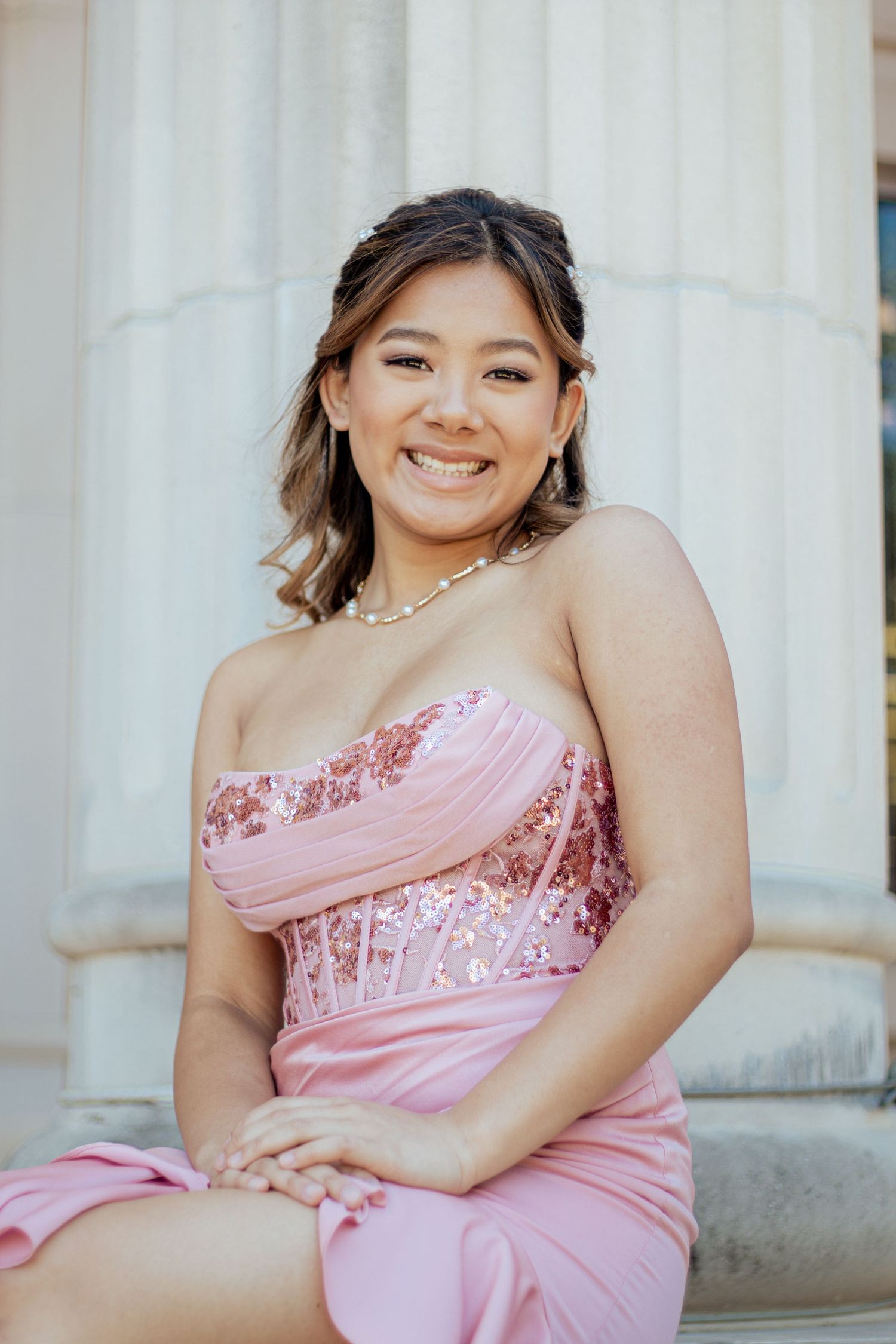Person in a pink strapless formal dress with ruched bodice posing in front of white stone columns.