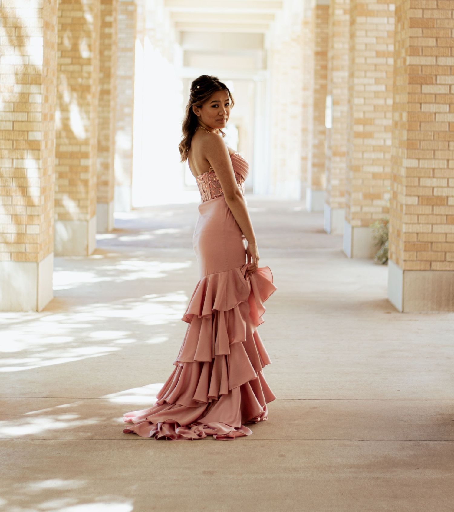 A figure in a flowing dusty rose gown with cascading ruffles stands in a sunlit columned hallway.