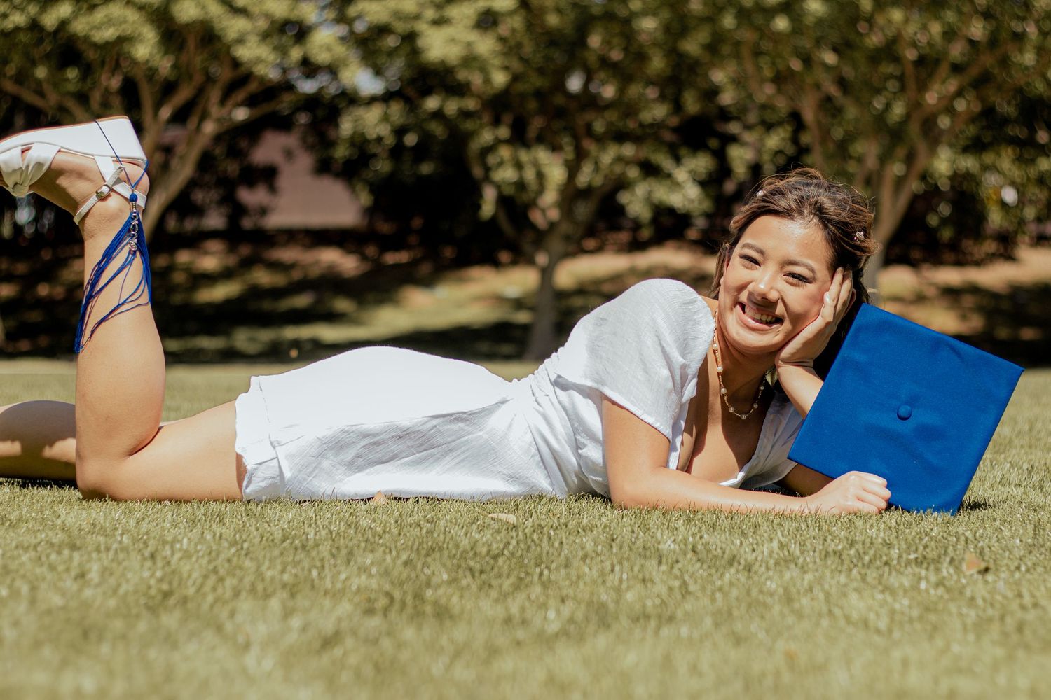 A person in white lounges on a blue blanket in the grass while reading outdoors on a sunny day.