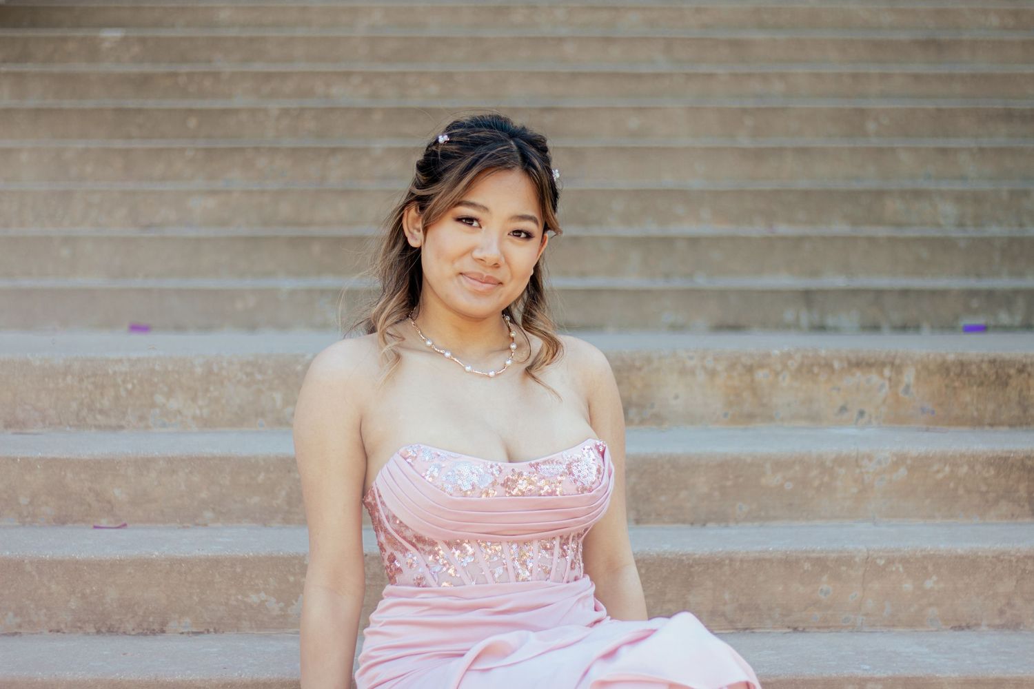 A person in a pink satin prom dress sits elegantly on outdoor stone steps.