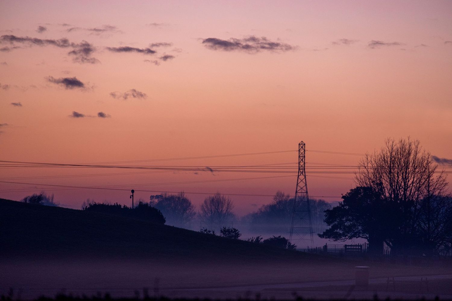Pastel sunrise over a misty rural landscape with silhouetted trees and a tall transmission tower in the distance.