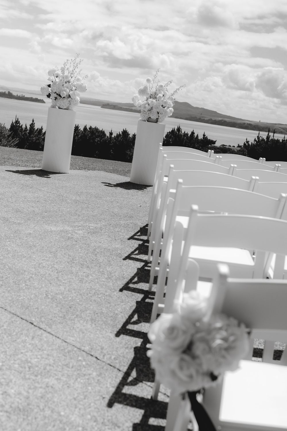 Black and white photo of white wedding chairs and floral arrangements set up for an outdoor ceremony with scenic views.
