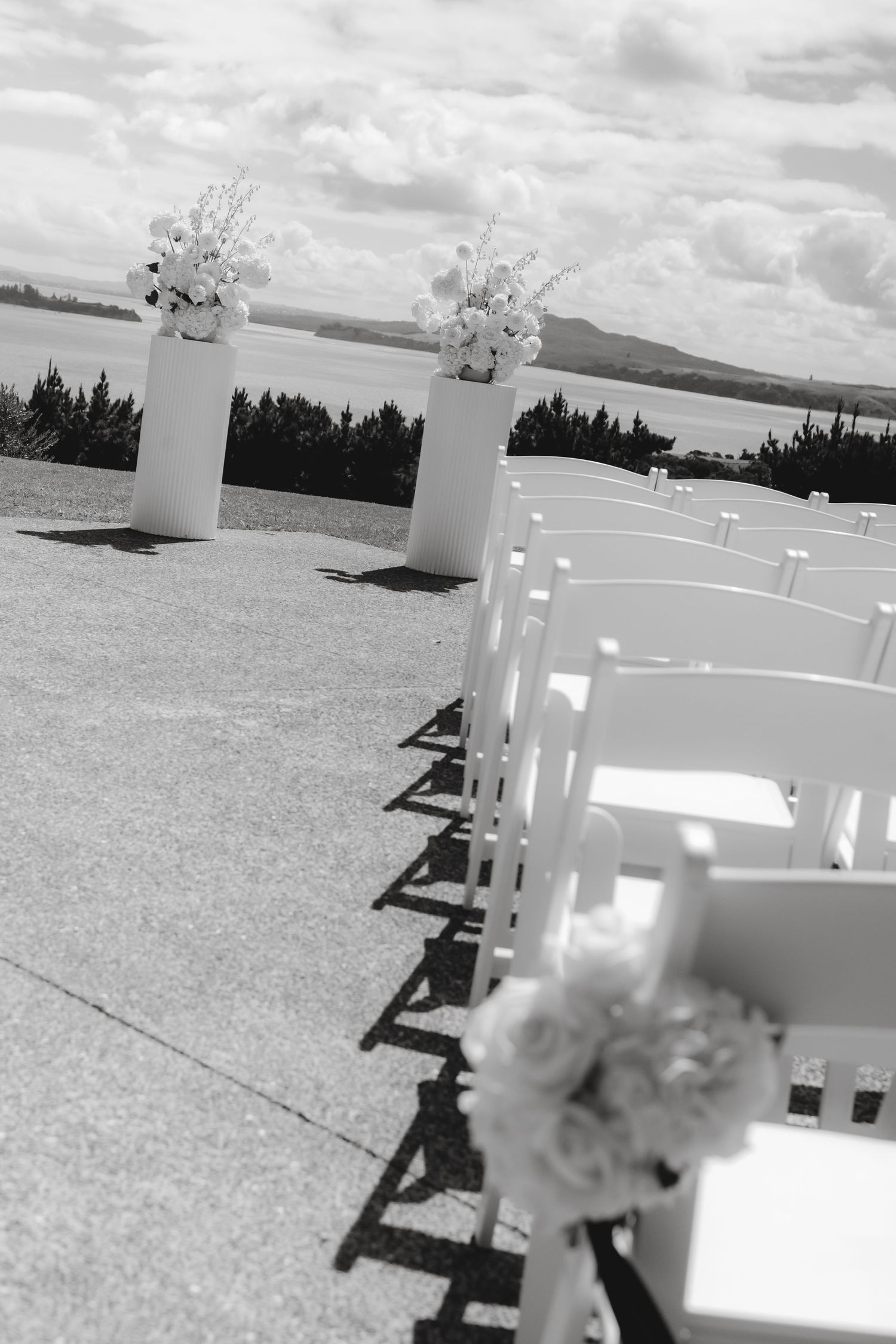 Black and white photo of white wedding chairs and floral arrangements set up for an outdoor ceremony with scenic views.