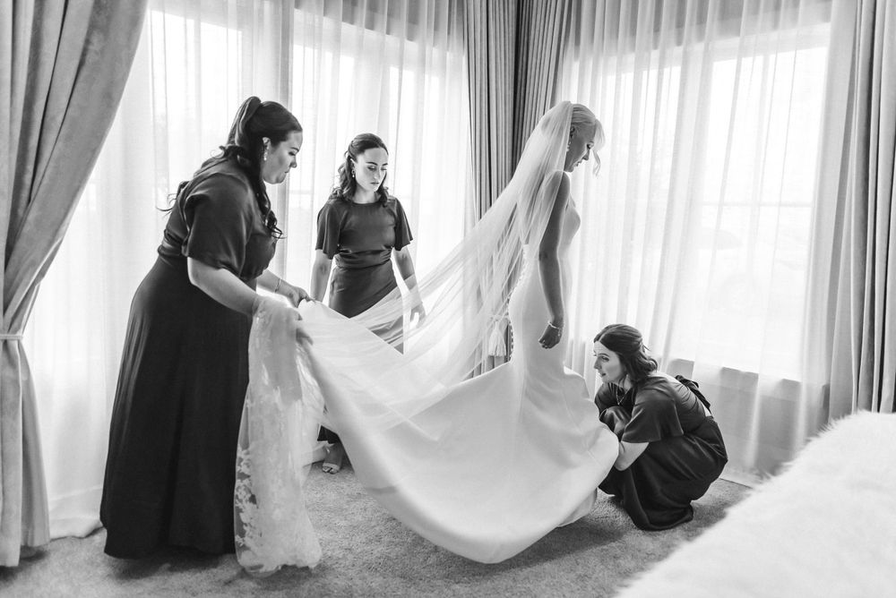 Black and white photo showing preparations before a wedding as attendants help adjust a long flowing veil.