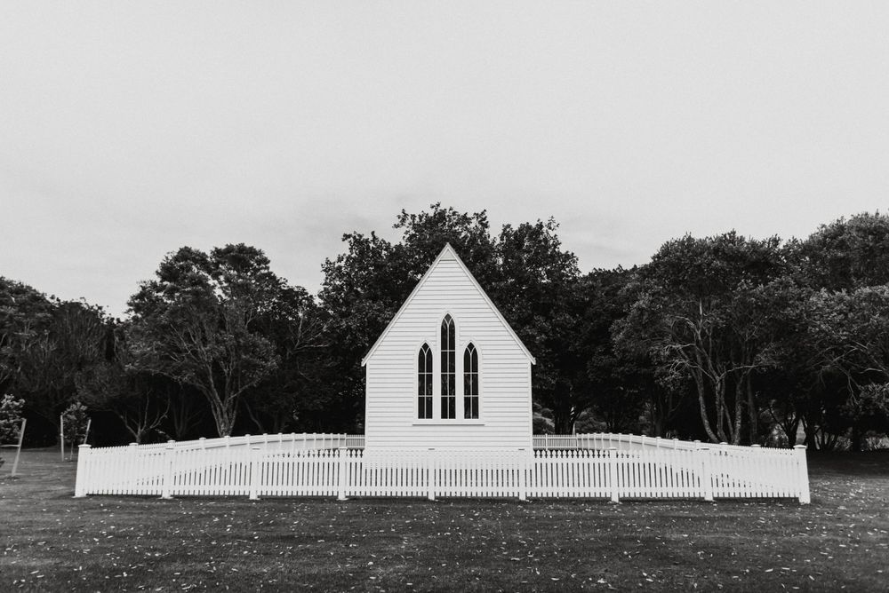 Small white chapel with gothic windows and picket fence stands alone against wooded background in black and white photo.