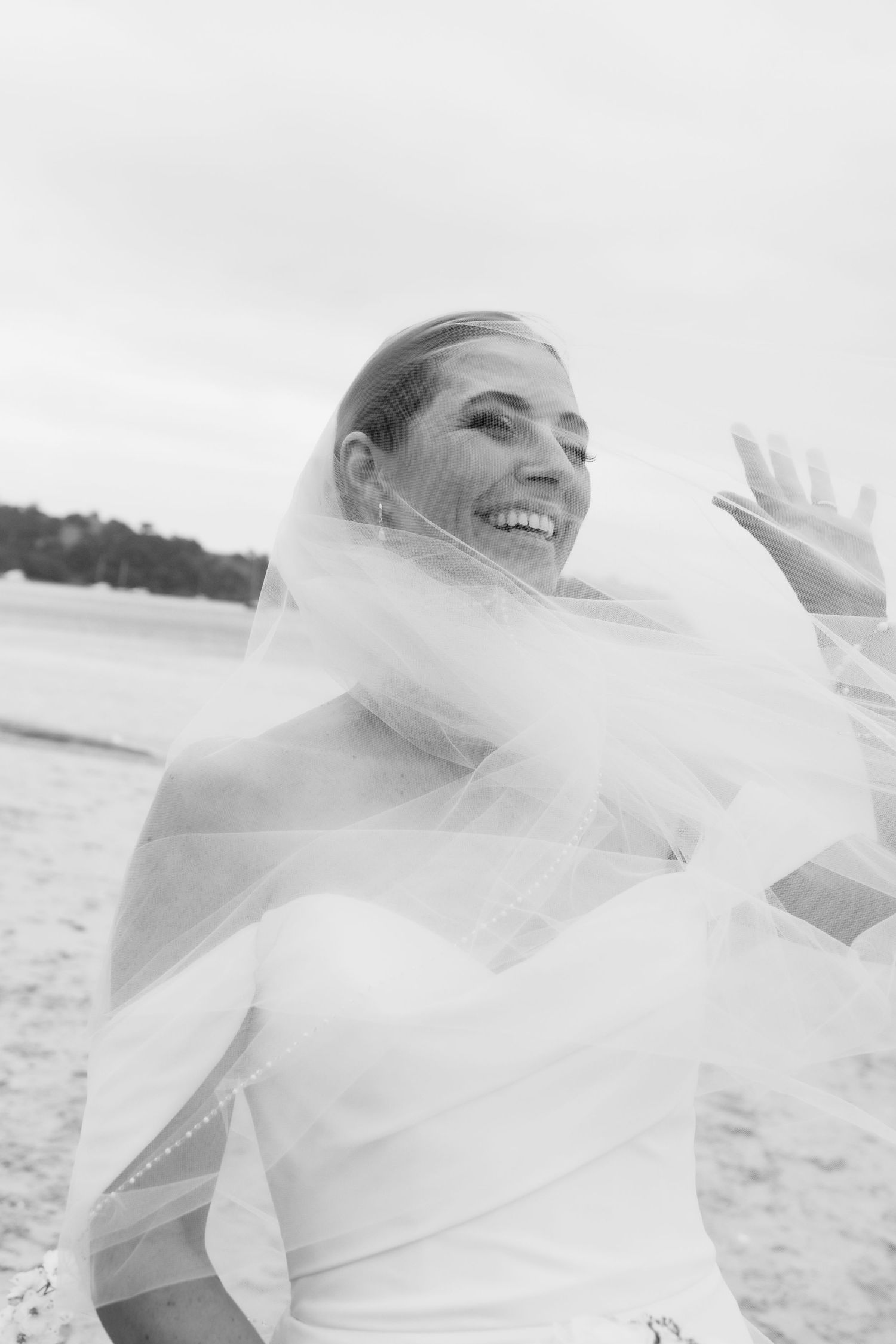 A joyful bride in a white wedding dress and veil smiles while standing on a beach in black and white photography.