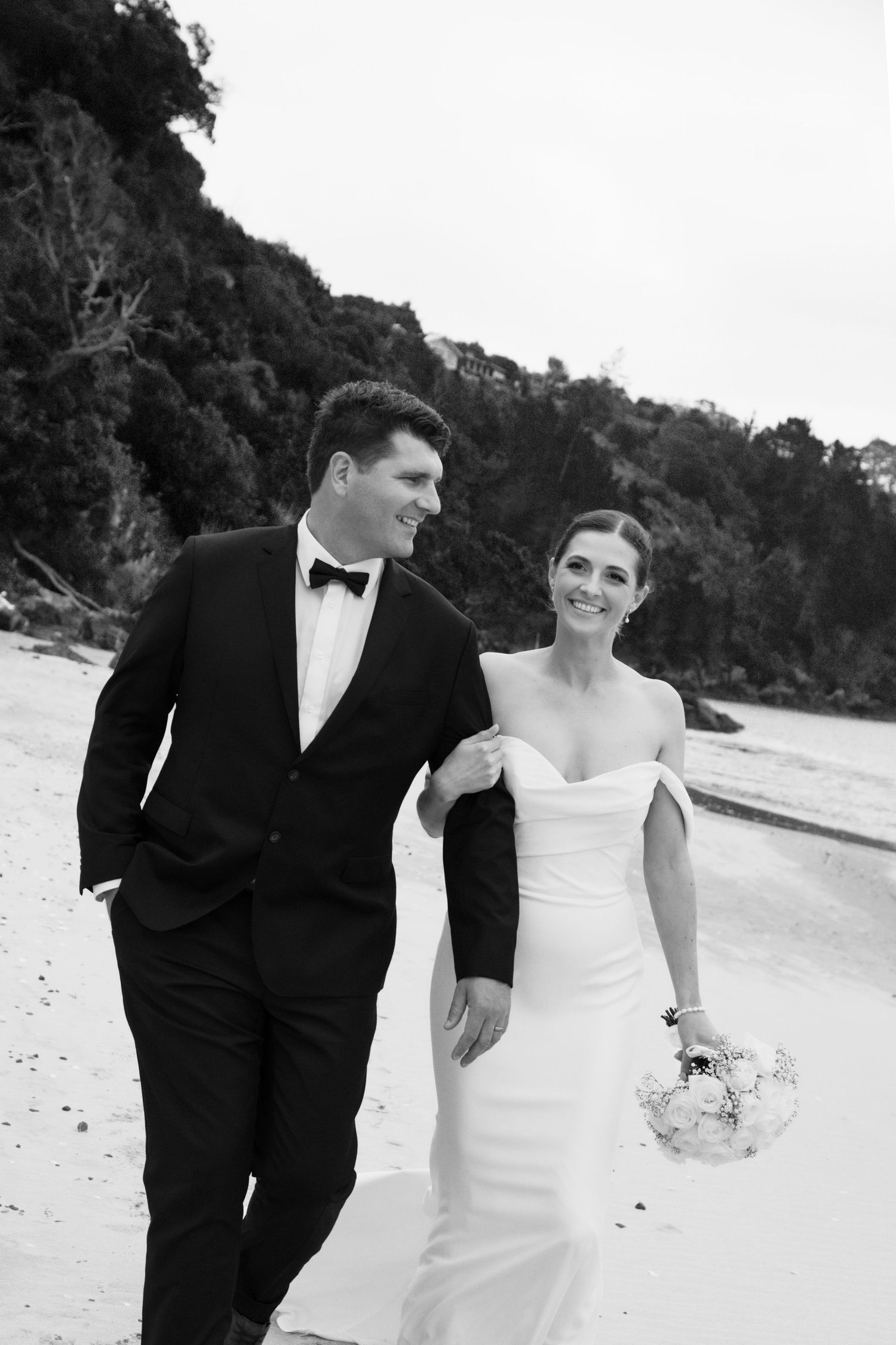 A couple in wedding attire walk together and smile on a beach with hills in the background.