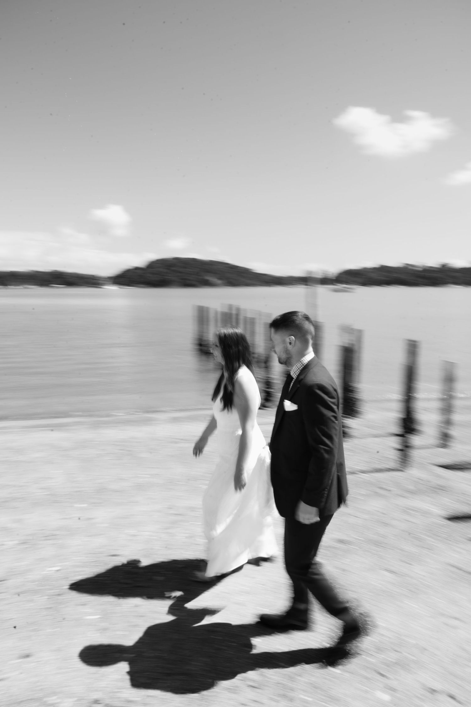 Black and white wedding photo showing couple walking on a dock by a lake with mountains in the background.