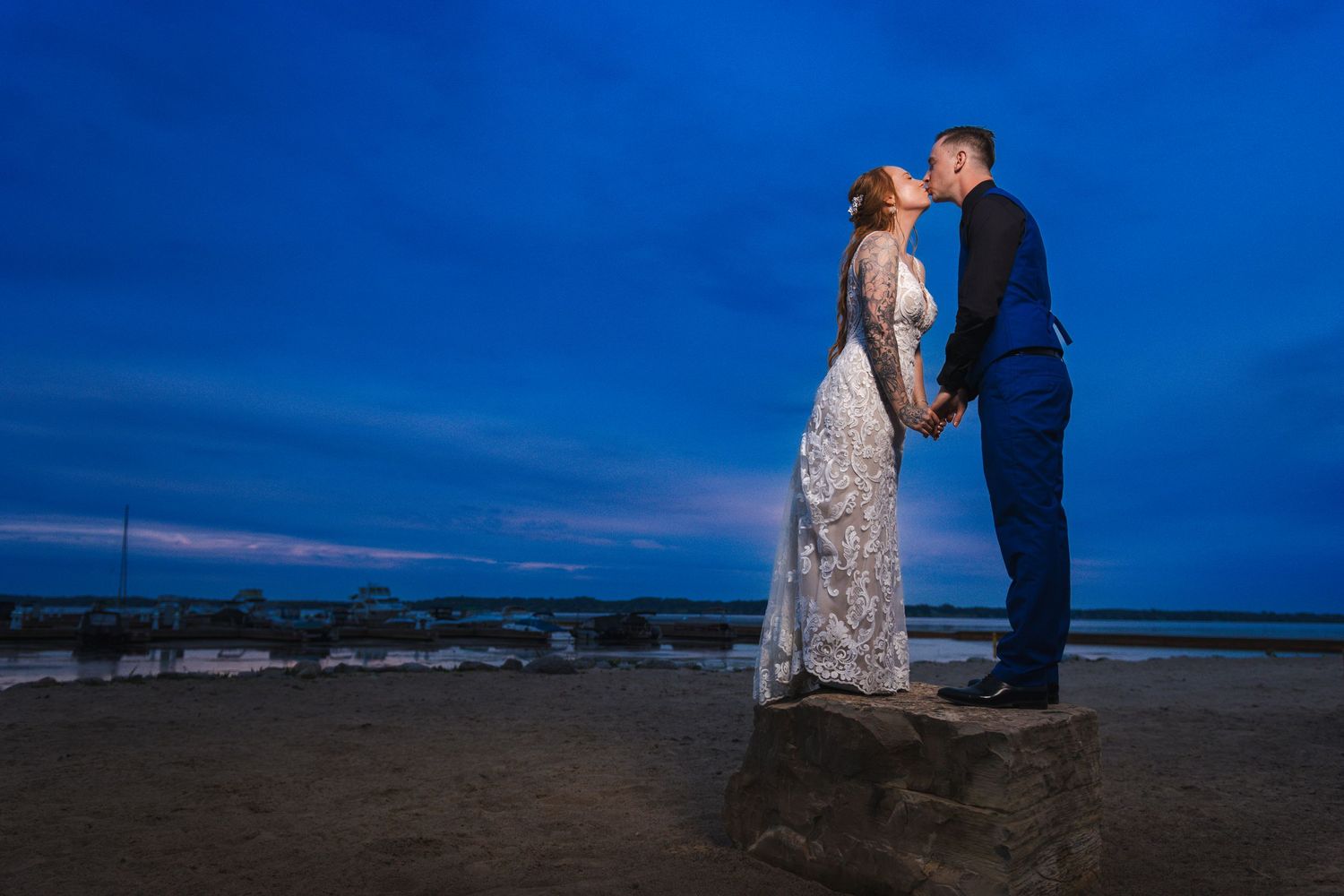 Two figures in wedding attire share a romantic moment at dusk against a dramatic deep blue sky on a beach.