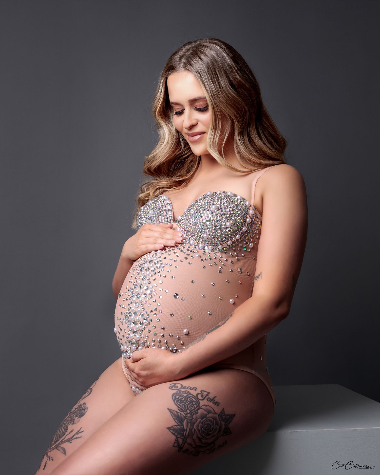 Mum in a sparkly bodysuit looking down lovingly at her belly, seated pose in studio.