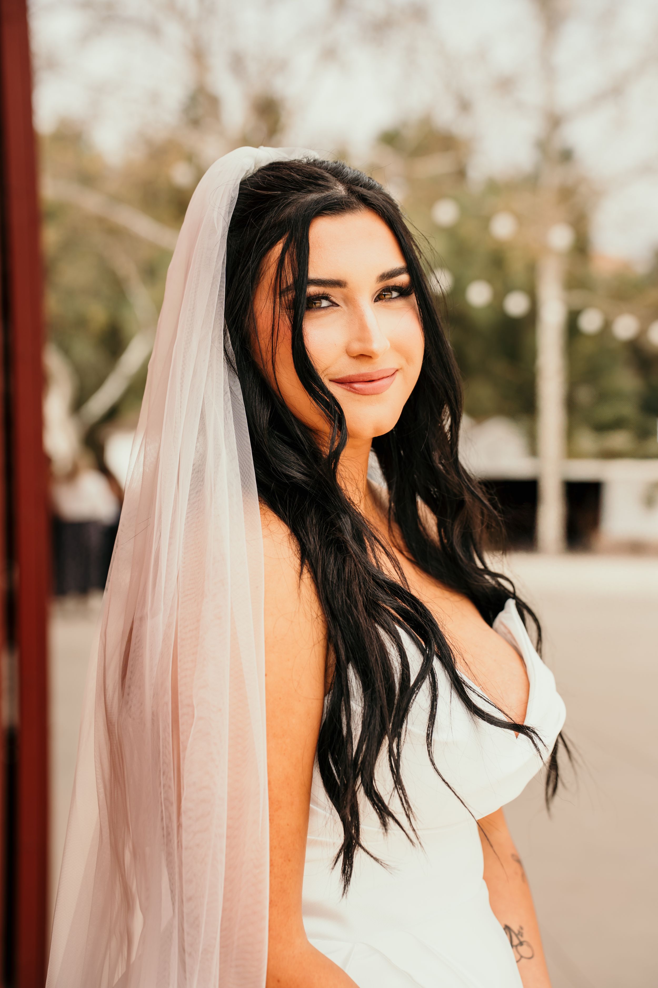 A smiling bride in a white wedding dress wears a flowing veil with long dark hair styled in loose waves.