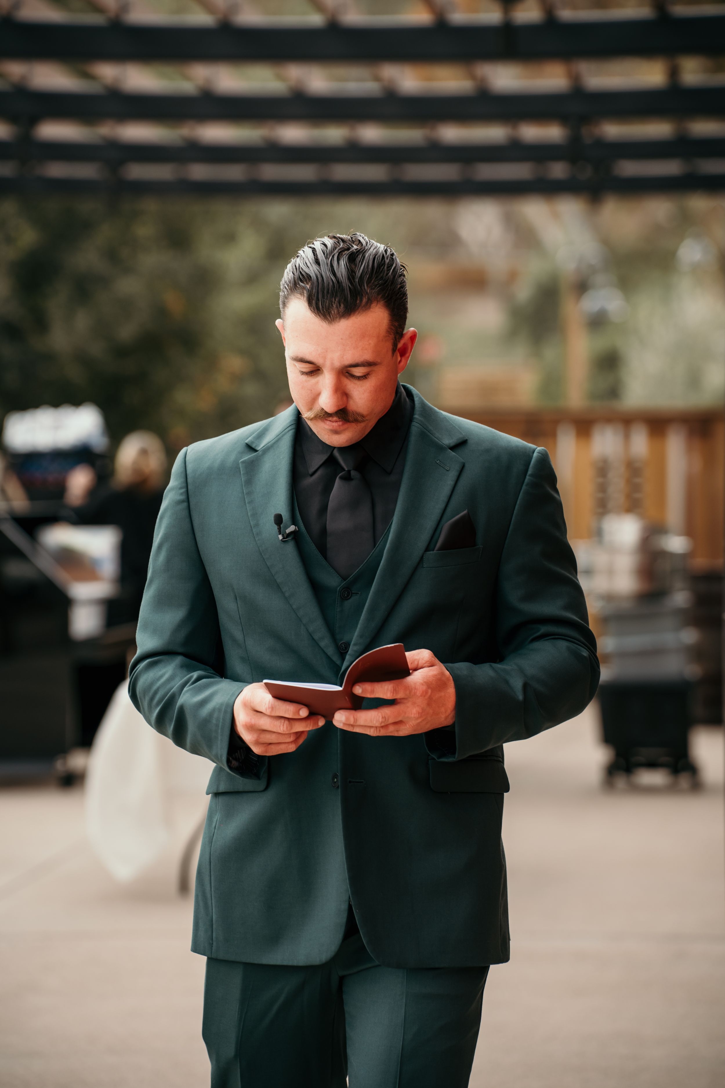 Fashionably dressed person in dark green suit checking phone while standing outdoors near wooden pergola.