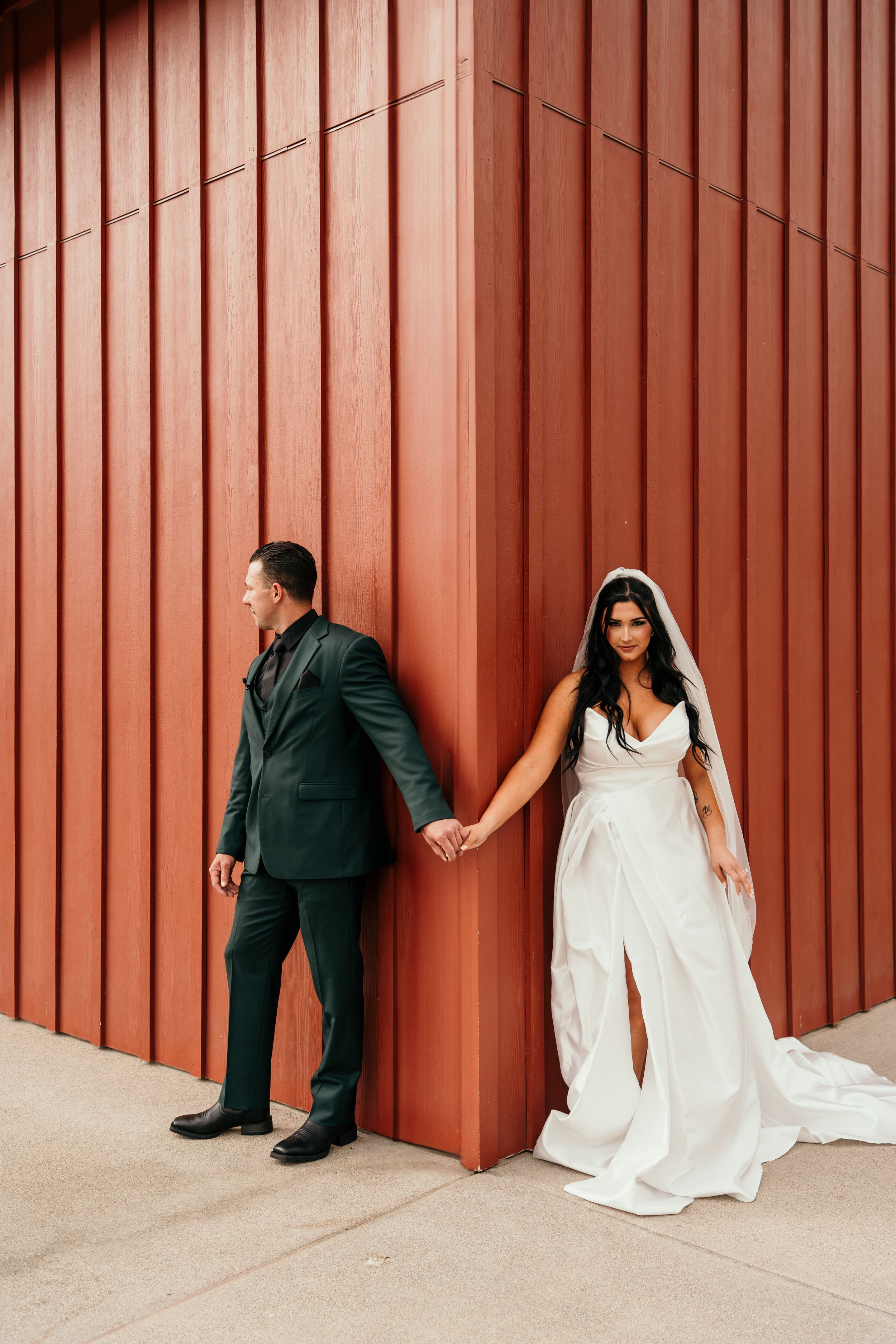 Bride in flowing white dress and groom in black suit hold hands while standing against rustic red wall.