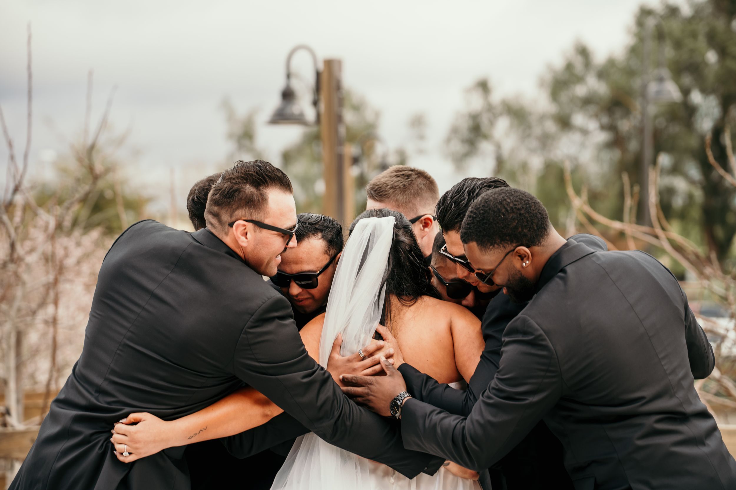 A group in formal attire shares an intimate group hug outdoors during what appears to be a wedding celebration.
