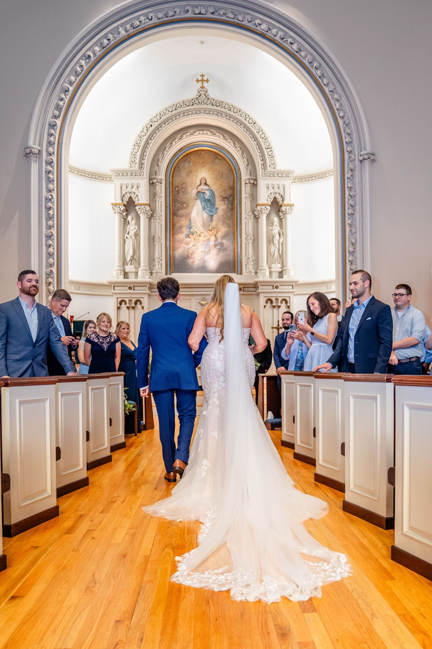 Couple walking down church aisle with ornate altar and religious artwork visible during wedding ceremony.