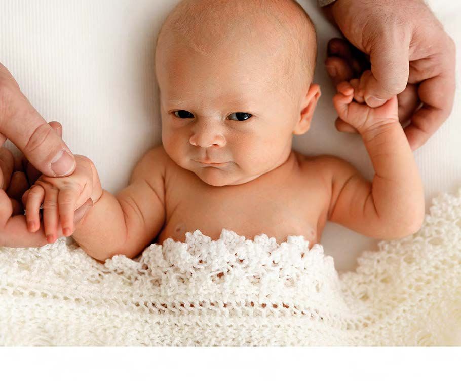 Infant lying on soft white crochet blanket while hands hold their tiny arms against delicate fabric.