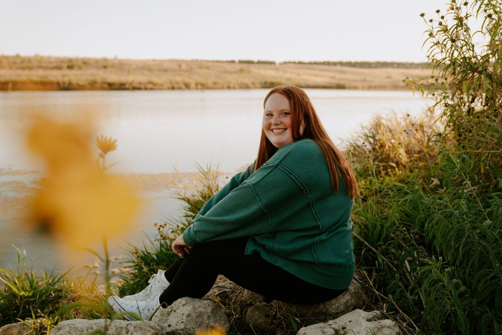 A person in a green sweater sits in a field during golden hour with tall grass and a warm sunset in the background.