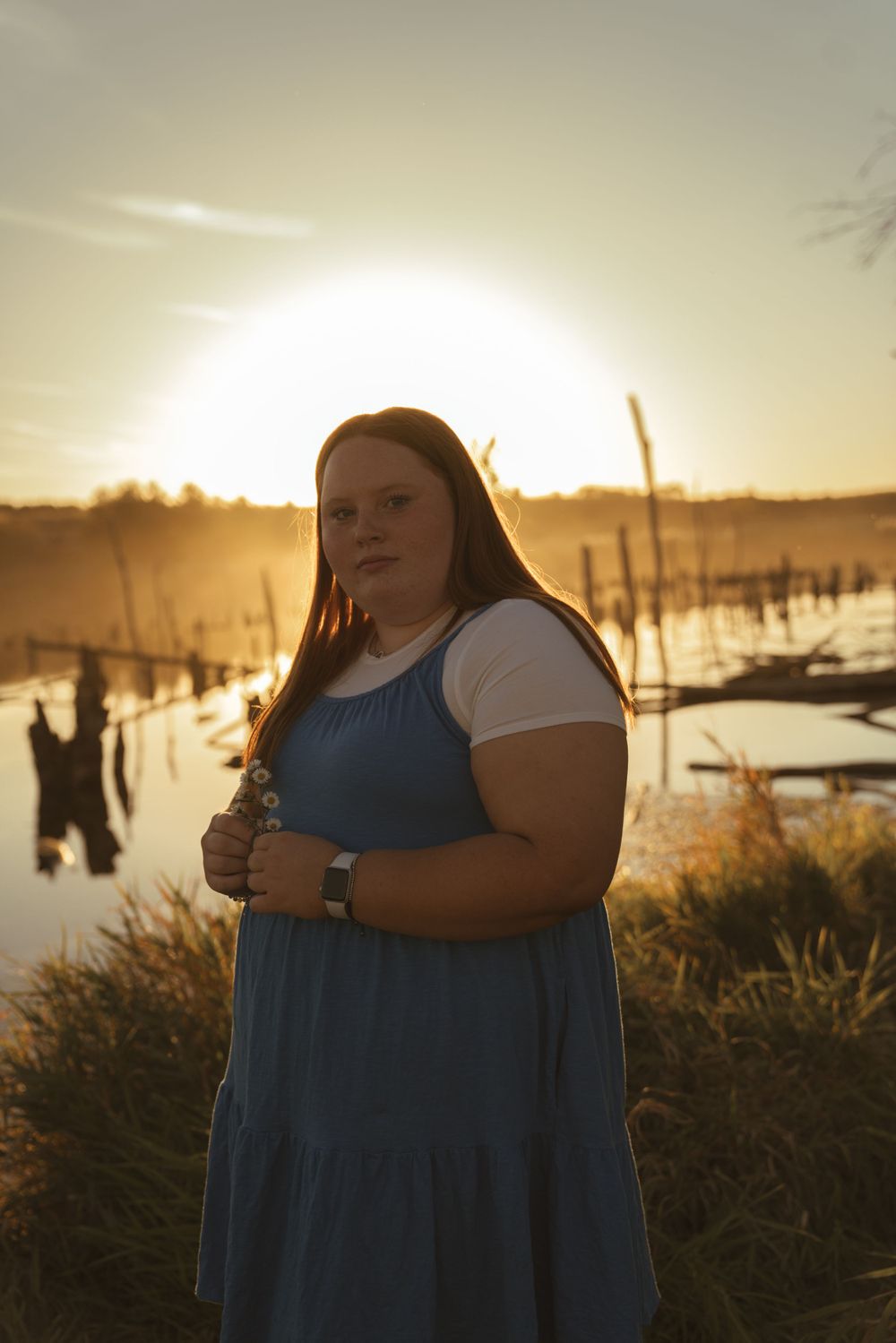 Figure in a blue dress poses against a sunset backdrop with water and wooden posts visible in the background.