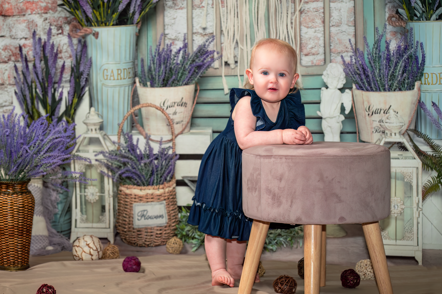 Small child in navy dress stands at stool surrounded by rustic decor and purple flowers in vintage setting.