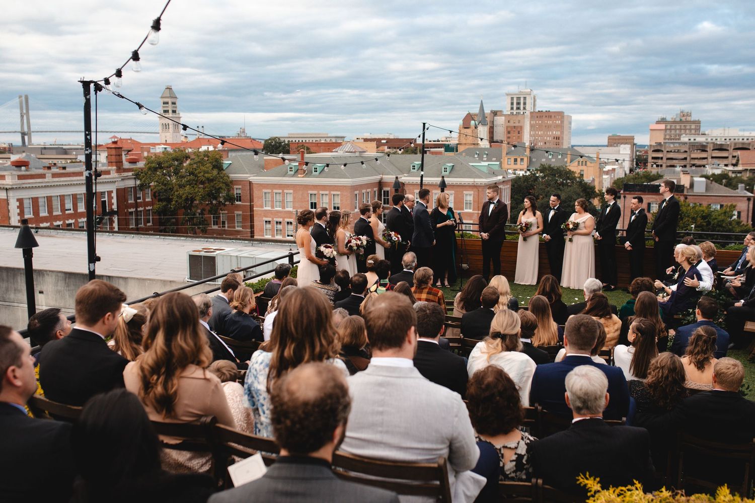 Rooftop wedding ceremony with city skyline backdrop, guests seated outdoors under string lights.