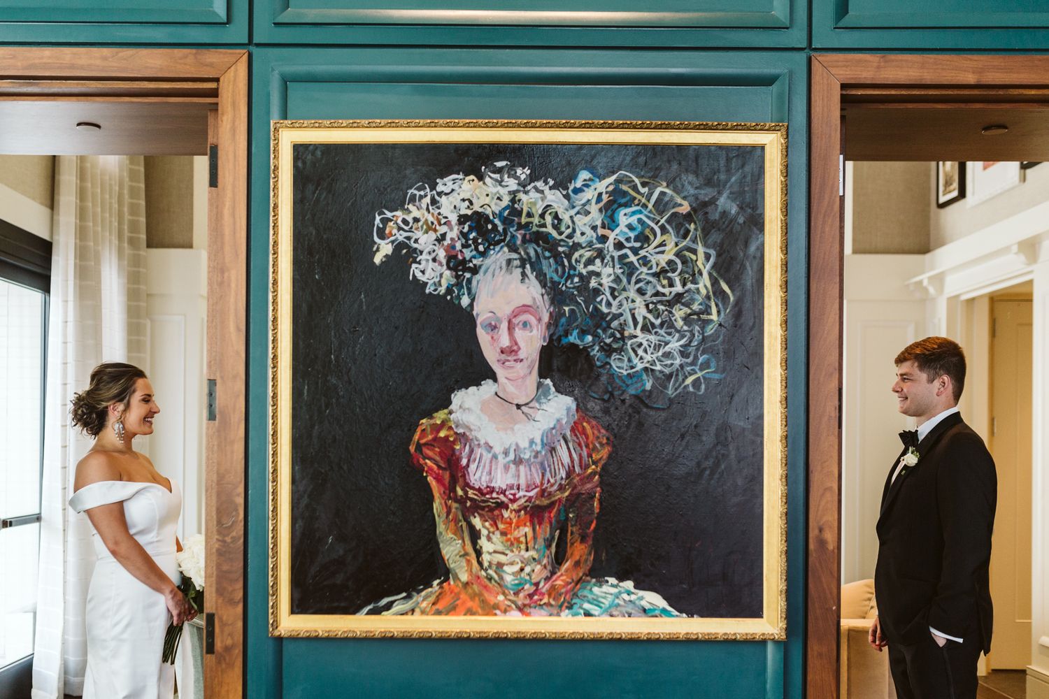 Bride and groom stand beside a large ornate portrait painting in a teal-walled venue during their wedding.