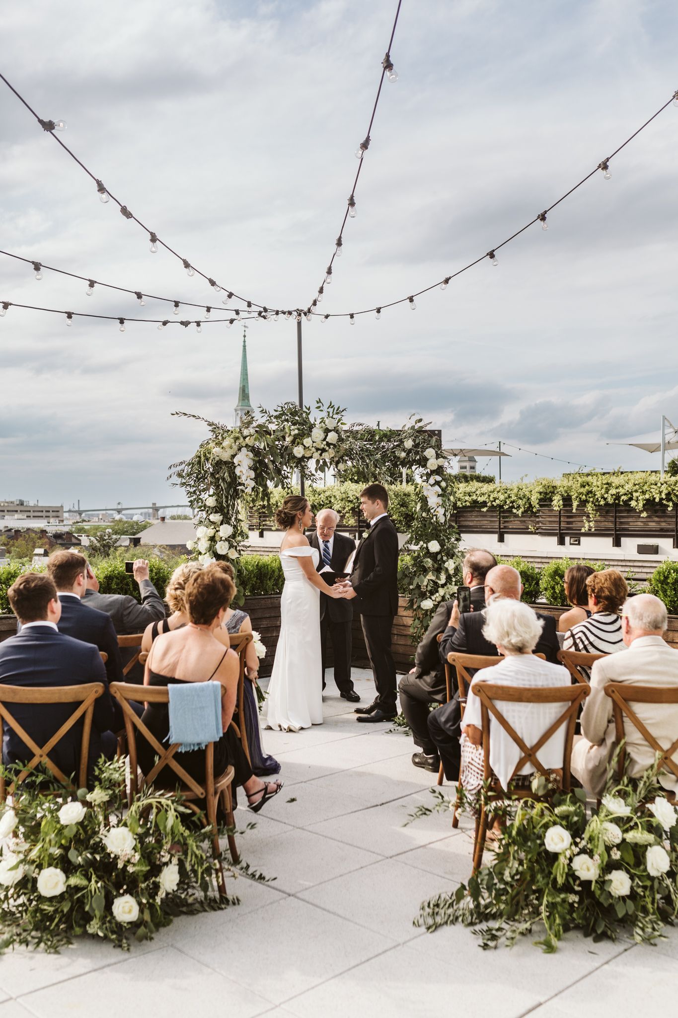 Rooftop wedding ceremony with floral arch, string lights, and guests seated on wooden chairs overlooking a city skyline.