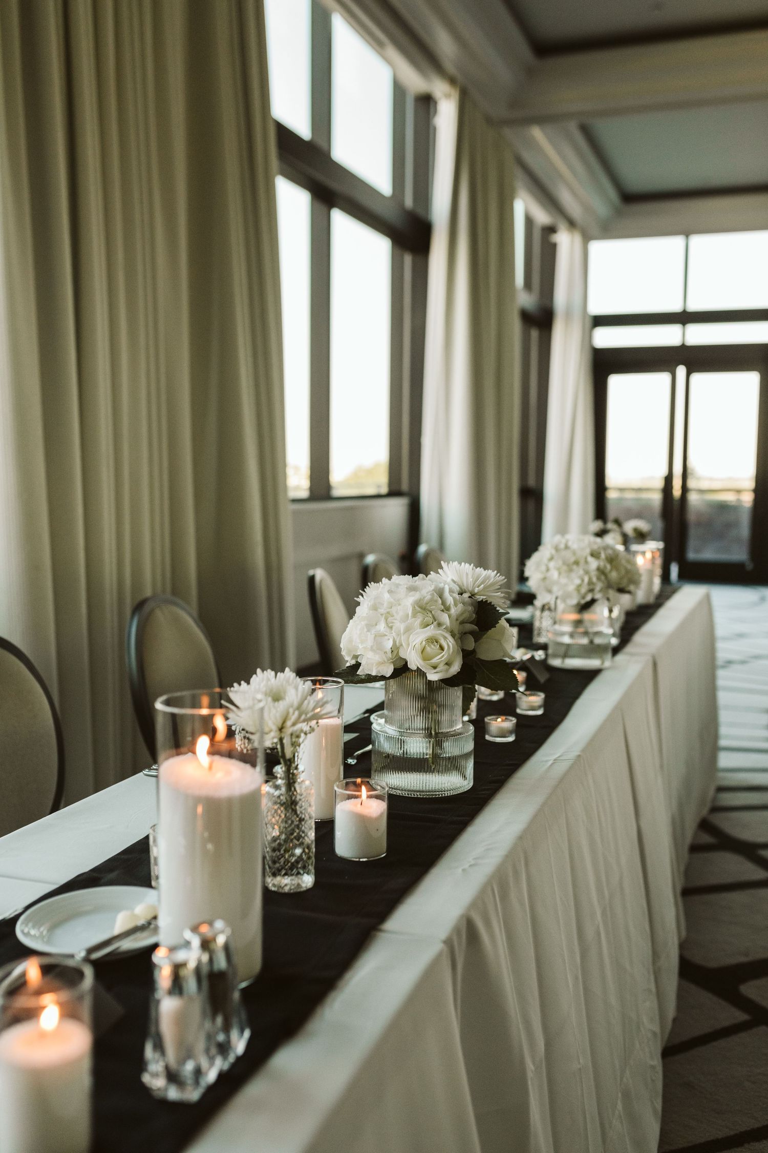 Elegant wedding head table with white floral centerpieces, lit pillar candles, and dark runner in a bright ballroom.