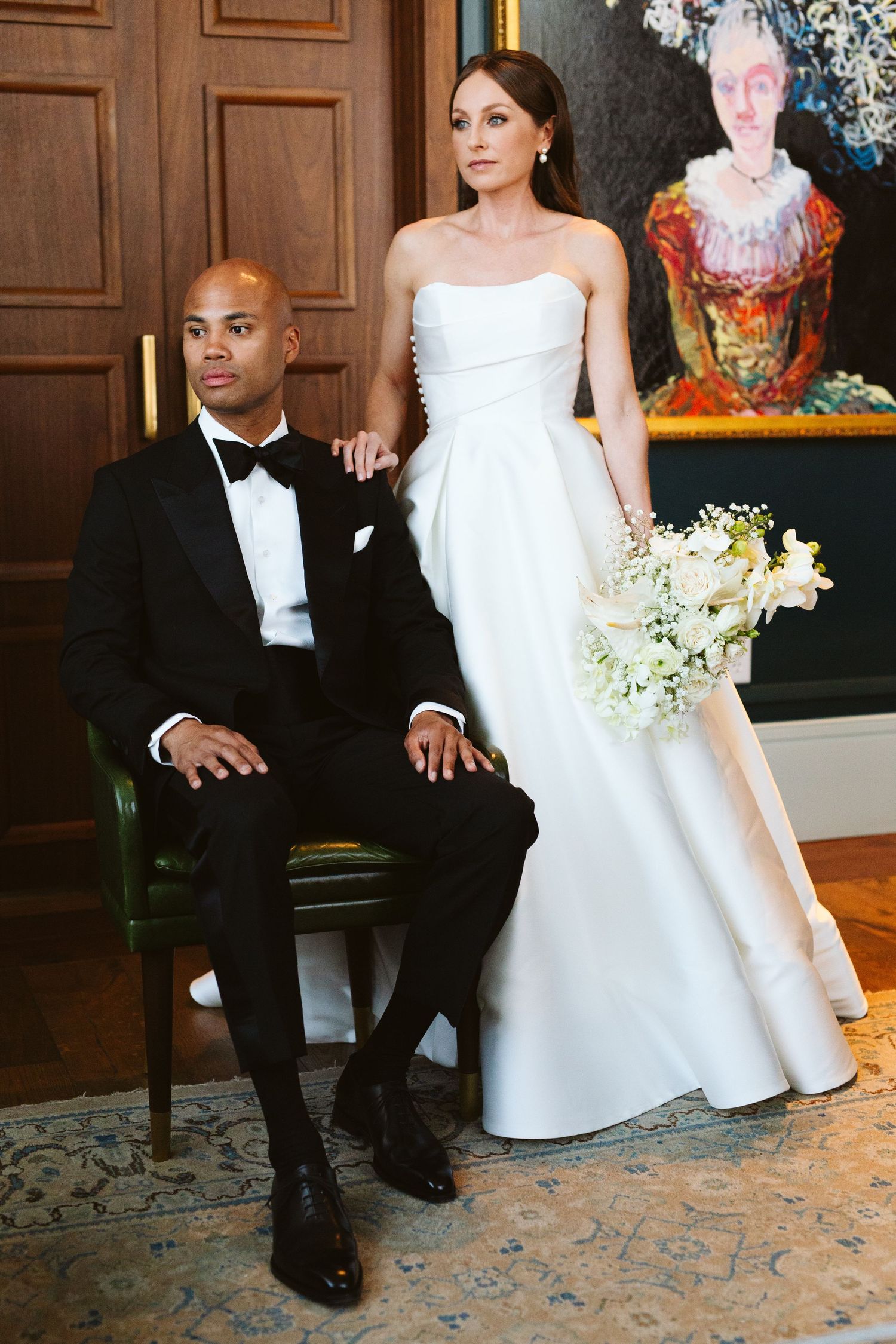 Elegant interracial wedding couple posing formally, groom in black tuxedo seated, bride standing in strapless white gown holding bouquet.