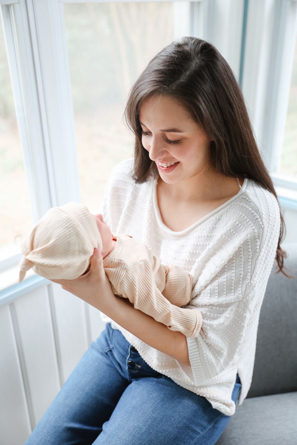 A smiling young mother with long dark hair holds her newborn baby wrapped in a blanket while sitting by a bright window.