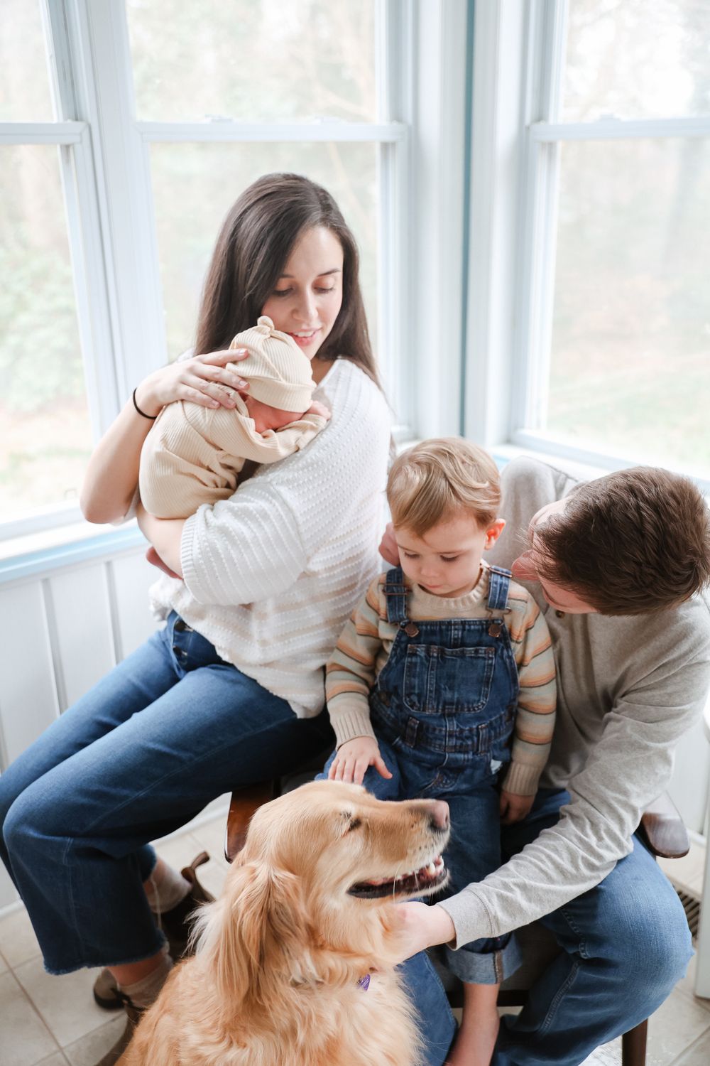 Happy family with newborn baby, toddler, and golden retriever dog sitting by bright windows.