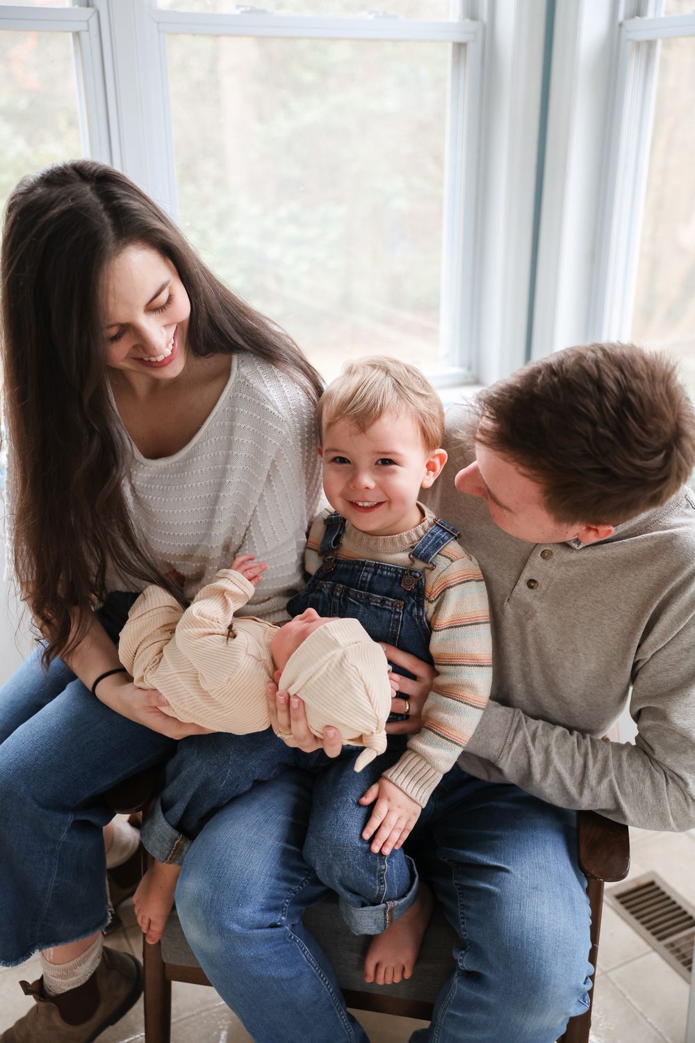 Happy young family with parents smiling at toddler holding newborn baby by bright window.