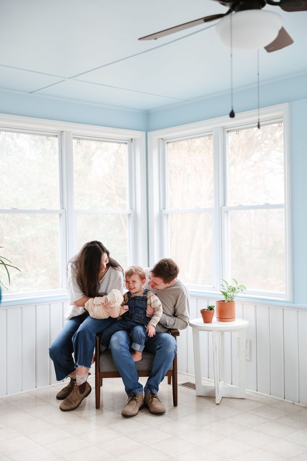 A young family sits together on a chair in a bright sunroom, parents lovingly attending to their toddler.