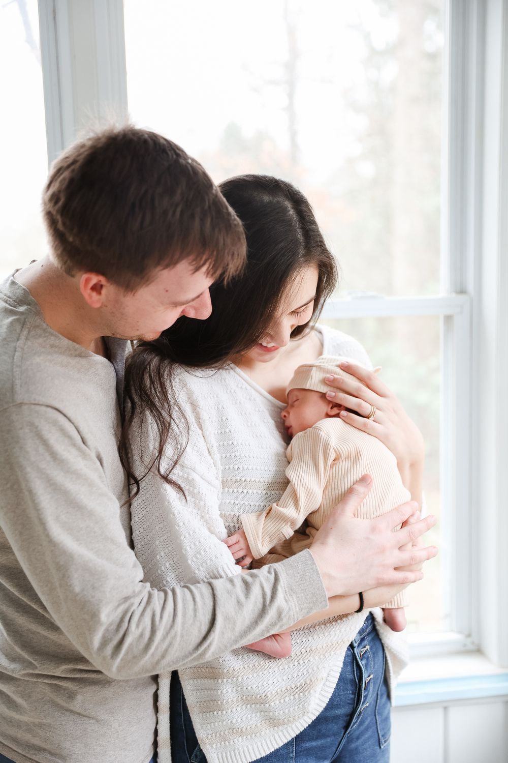 Young parents lovingly cradle their newborn baby together by a bright window at home.