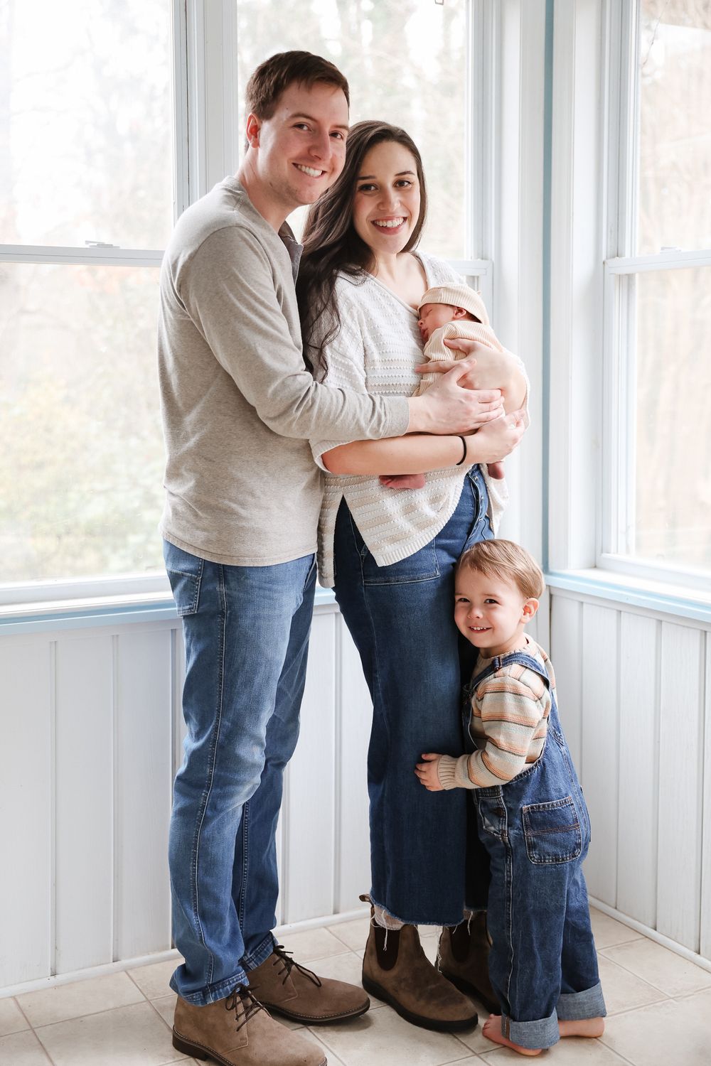 A happy young family of four poses indoors, parents holding a newborn baby while a toddler in overalls hugs mom's leg.