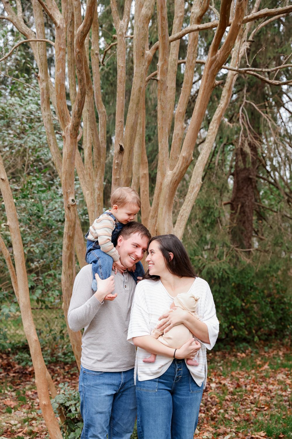 Happy family of three posing outdoors among trees, father carrying toddler on shoulders while mother holds newborn baby.