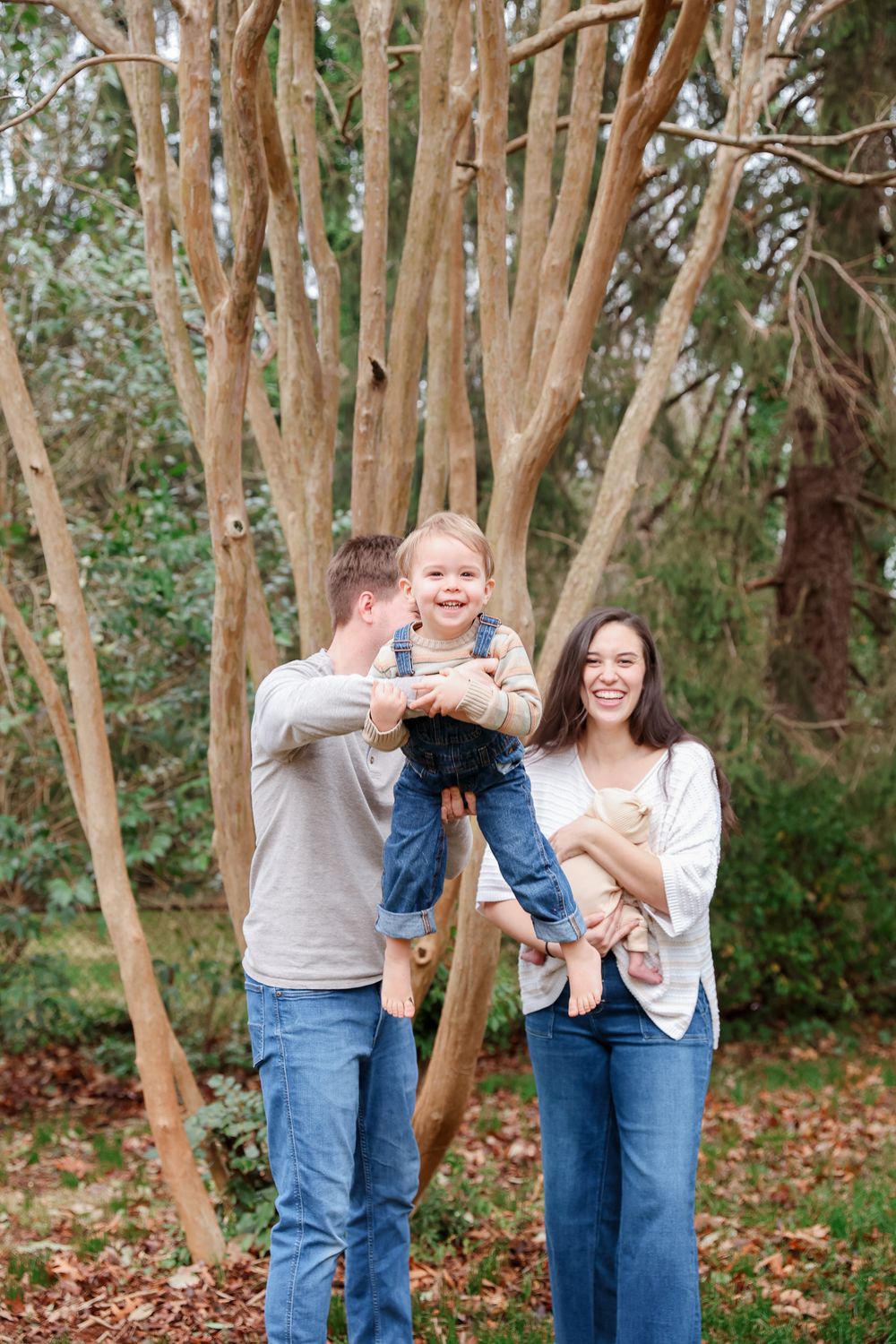 A happy young family with a toddler and baby poses outdoors among tall trees with autumn leaves on the ground.