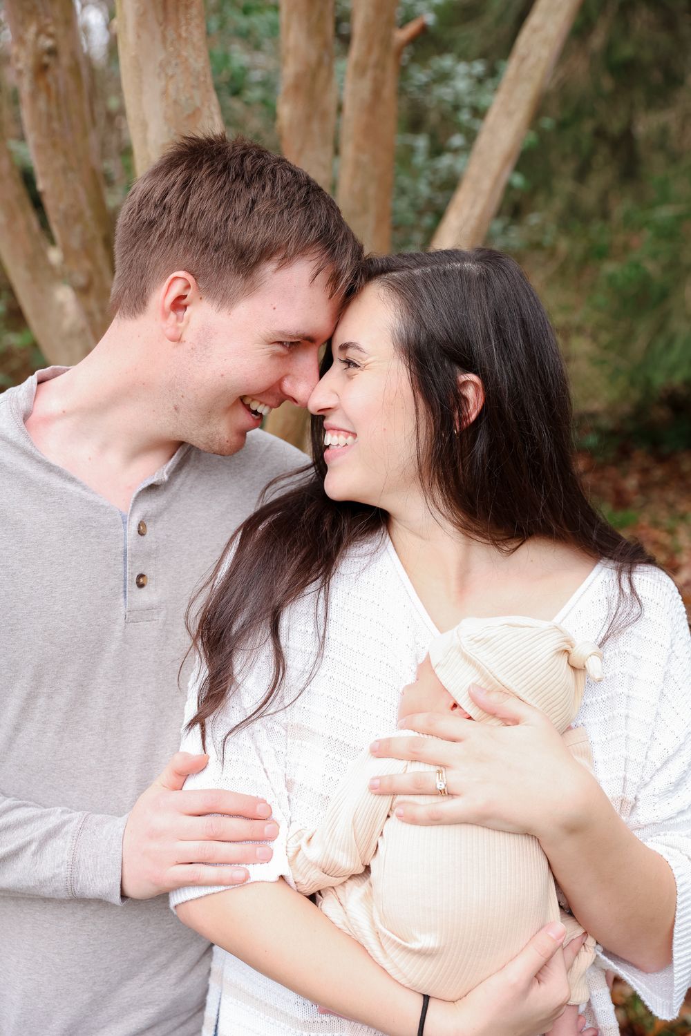 A happy couple smiles at each other while holding their newborn baby outdoors among trees.