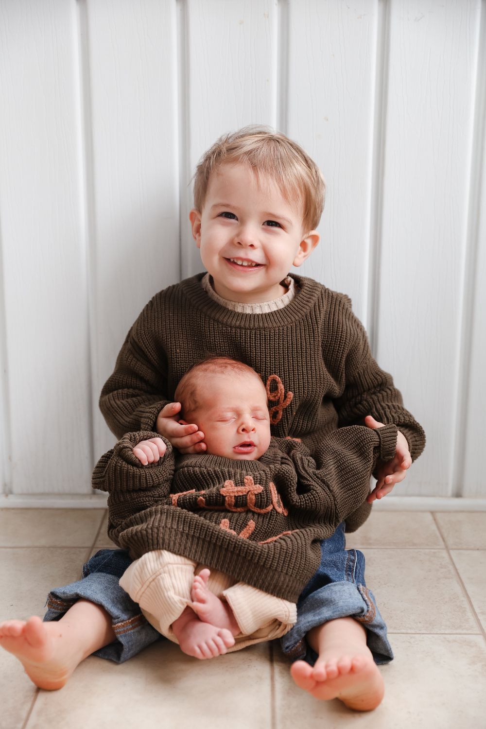 Smiling toddler boy in brown sweater gently holding sleeping newborn baby sibling.