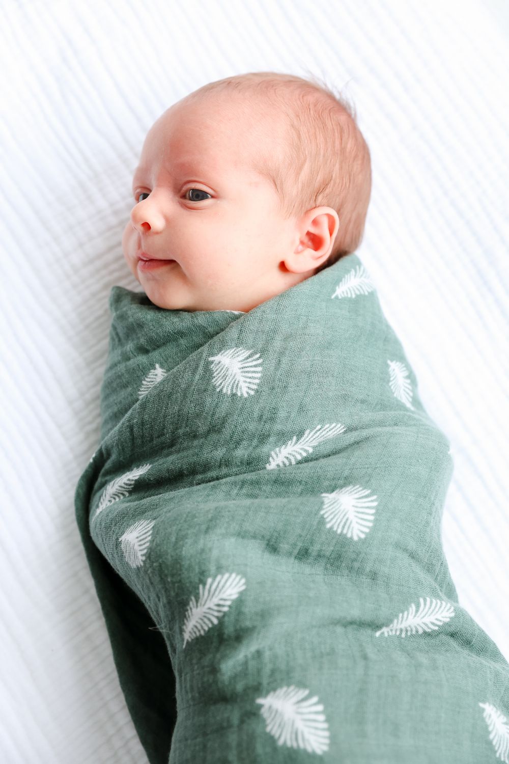 Peaceful newborn baby swaddled in green muslin blanket with white leaf pattern lying on white surface.