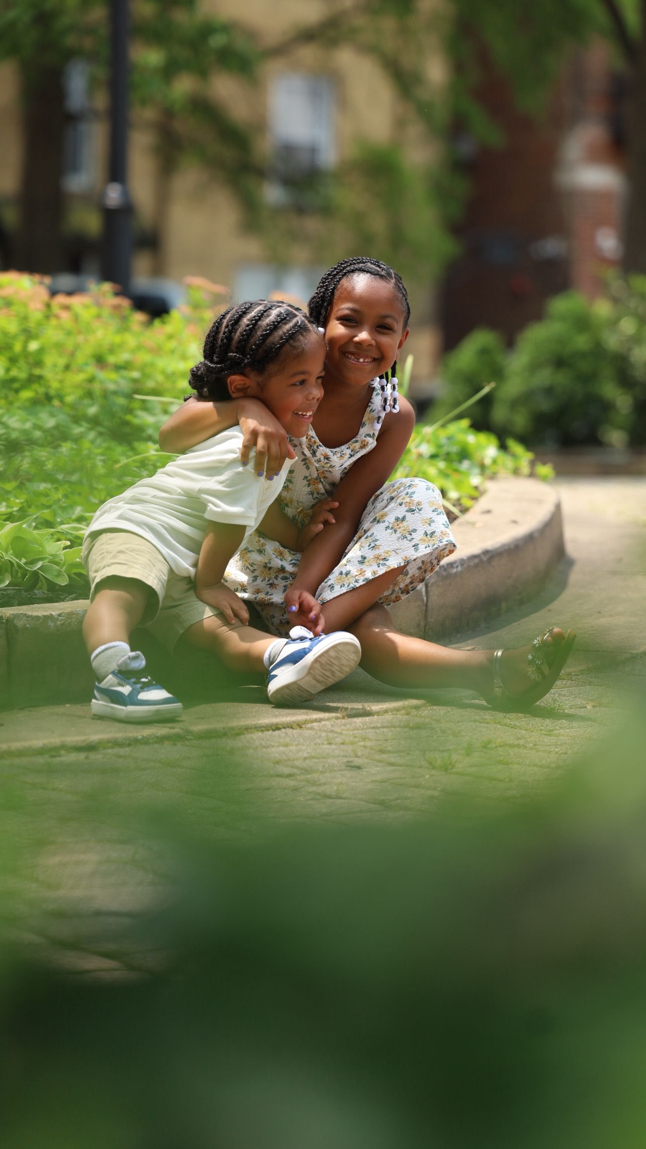 Two young Black children hugging and smiling while sitting outdoors in sunny park.
