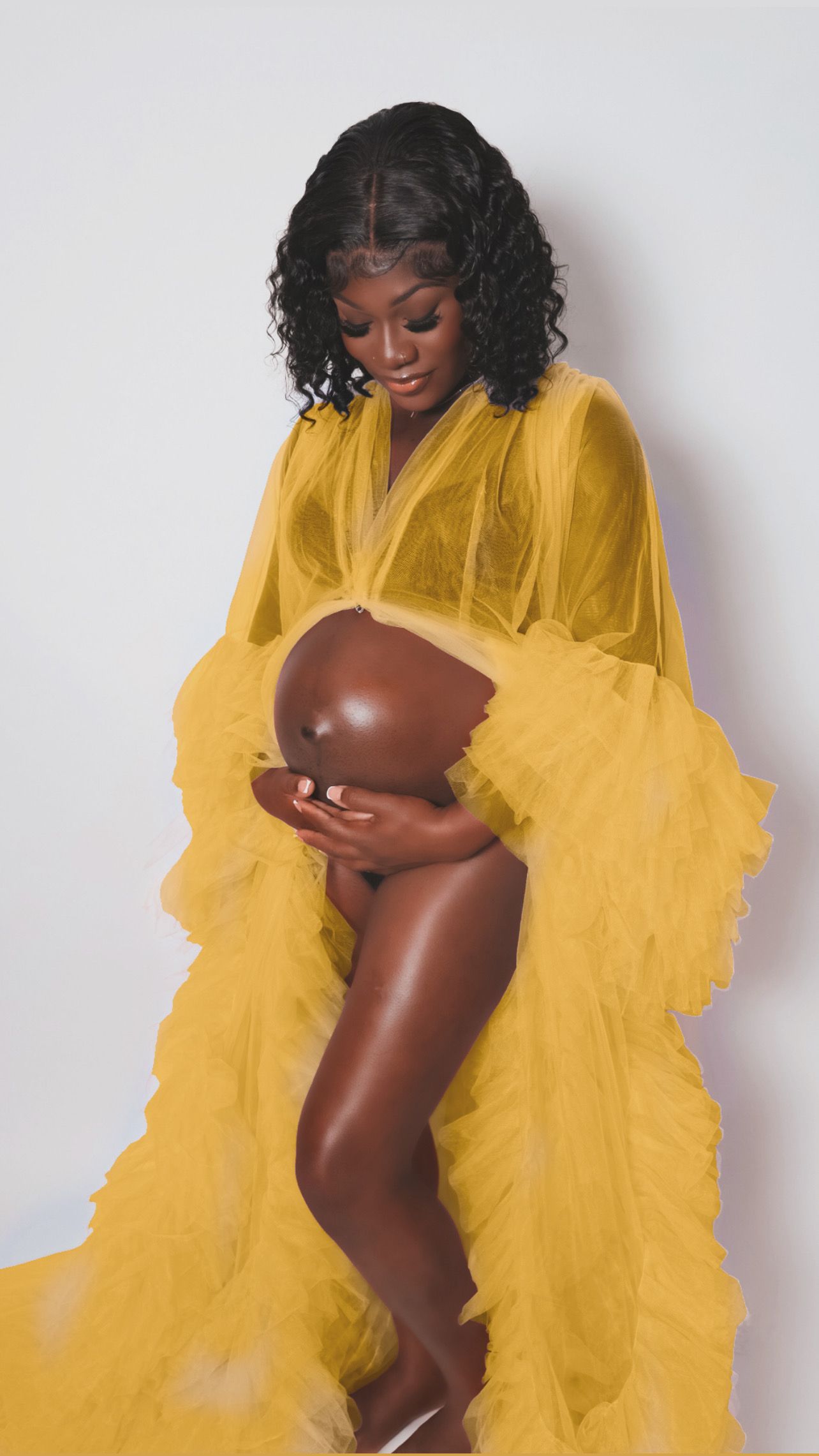 Pregnant Black woman in yellow tulle robe cradling baby bump in maternity portrait.