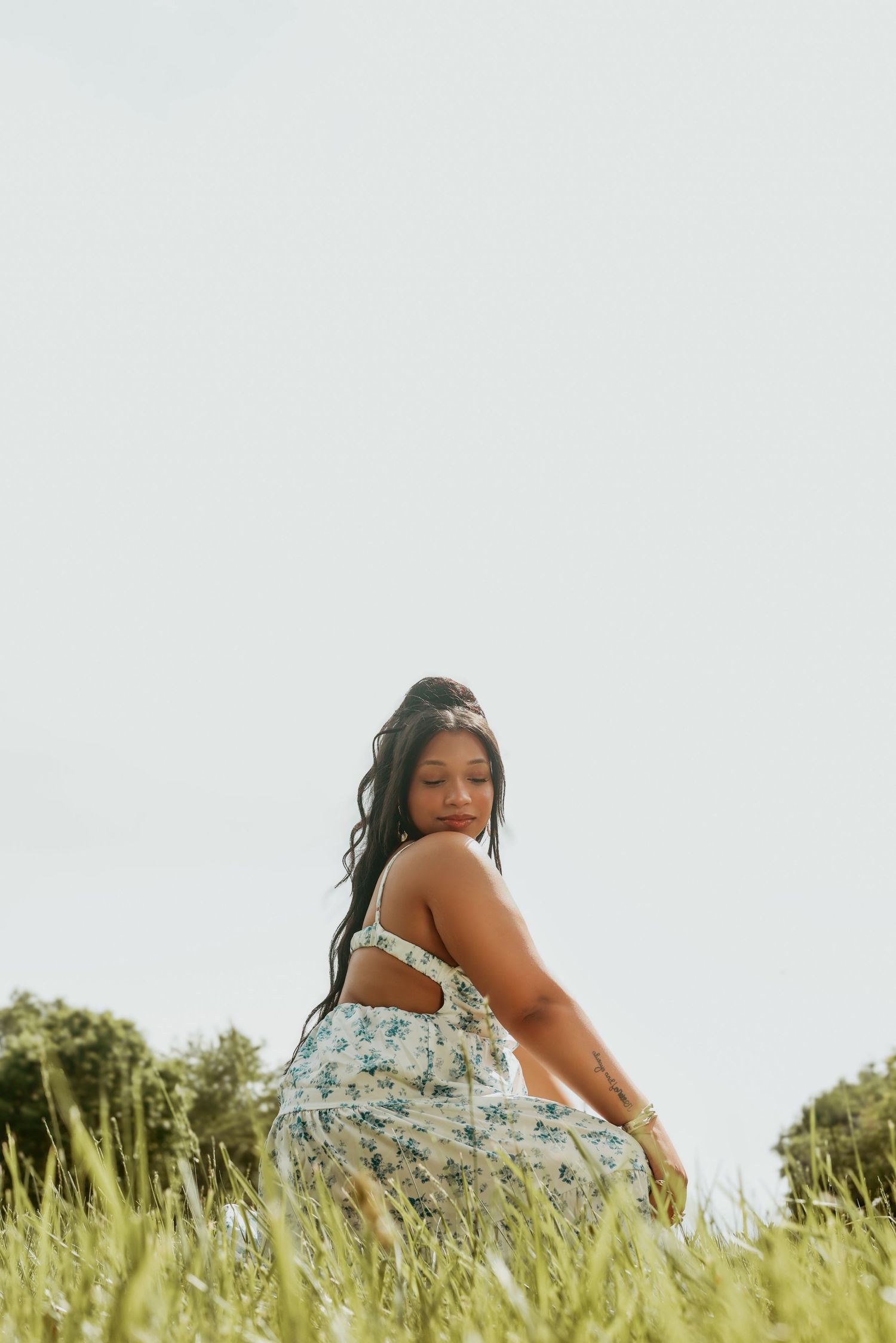 A young woman in a floral sundress sits in tall green grass beneath an overcast sky.