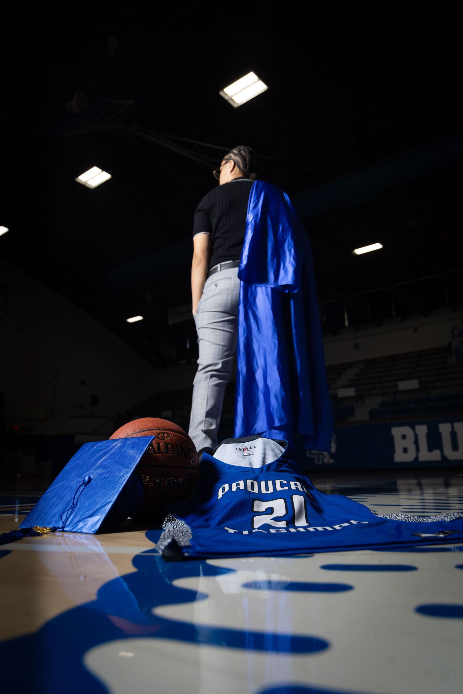 A student stands on a basketball court with blue graduation cap, gown, and jersey displayed.
