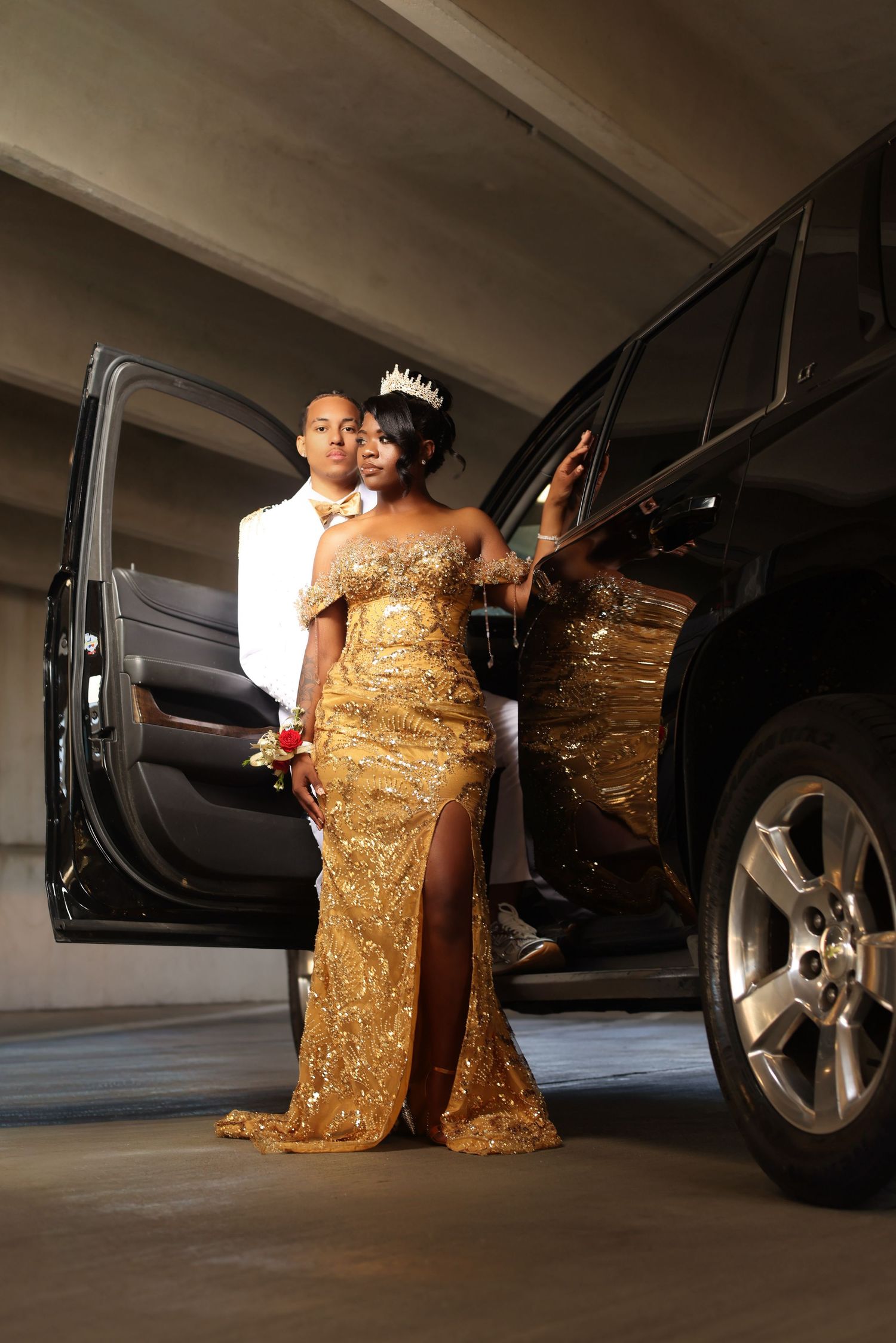A couple in formal attire poses by a black SUV, woman in gold sequin gown with crown.