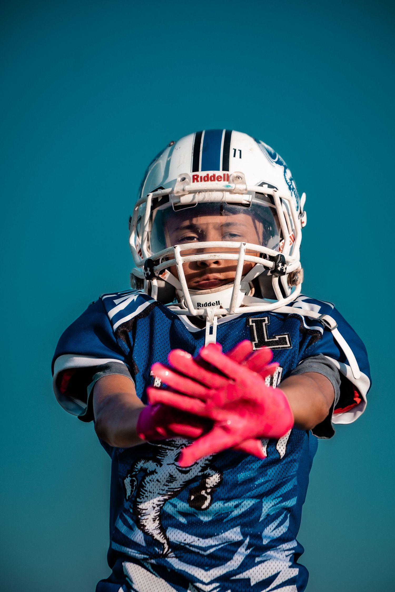 A young football player in a blue helmet and jersey reaches toward the camera with pink gloves.
