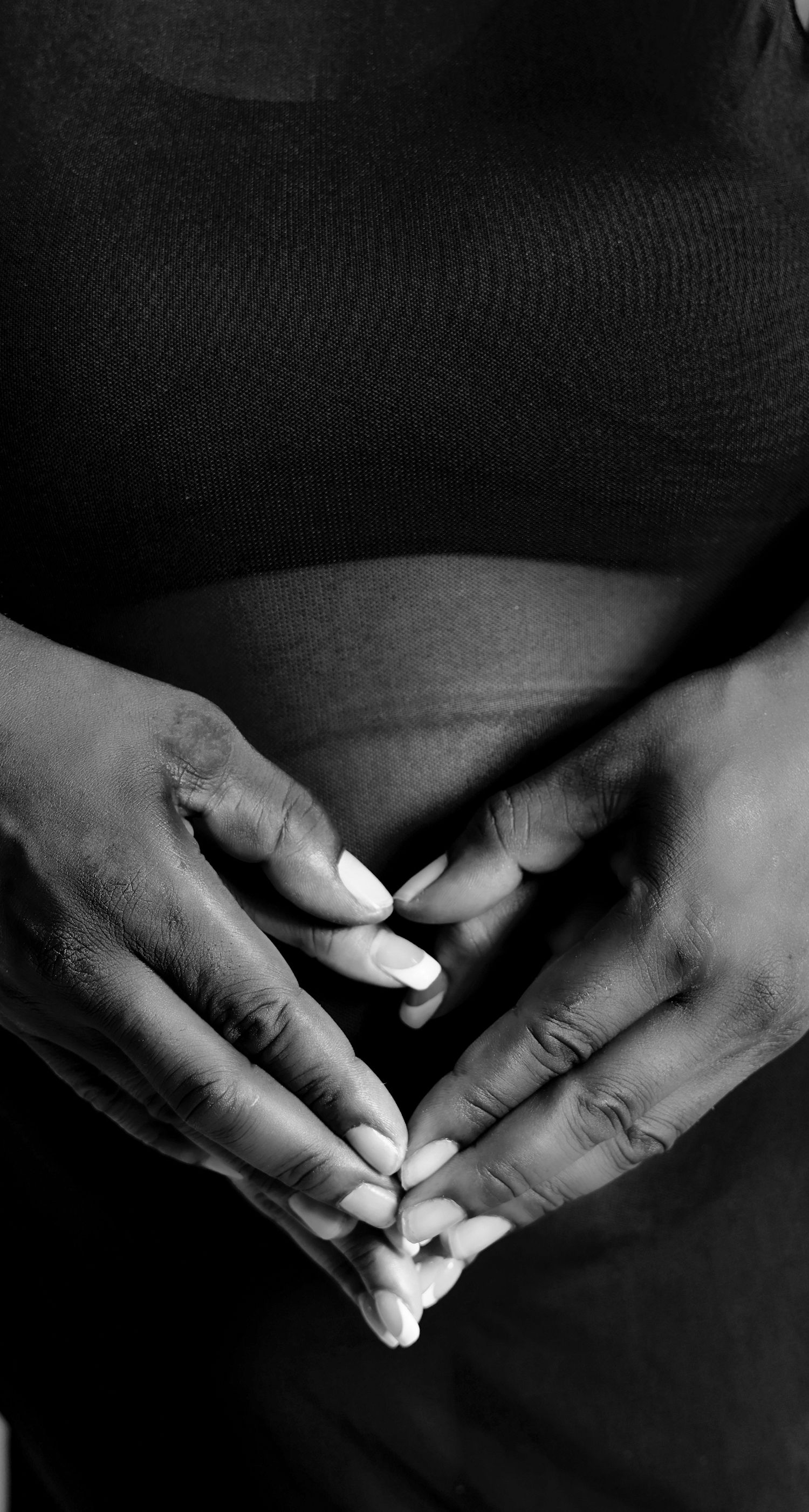 A black and white close-up of pregnant woman cradling her baby bump with manicured hands.